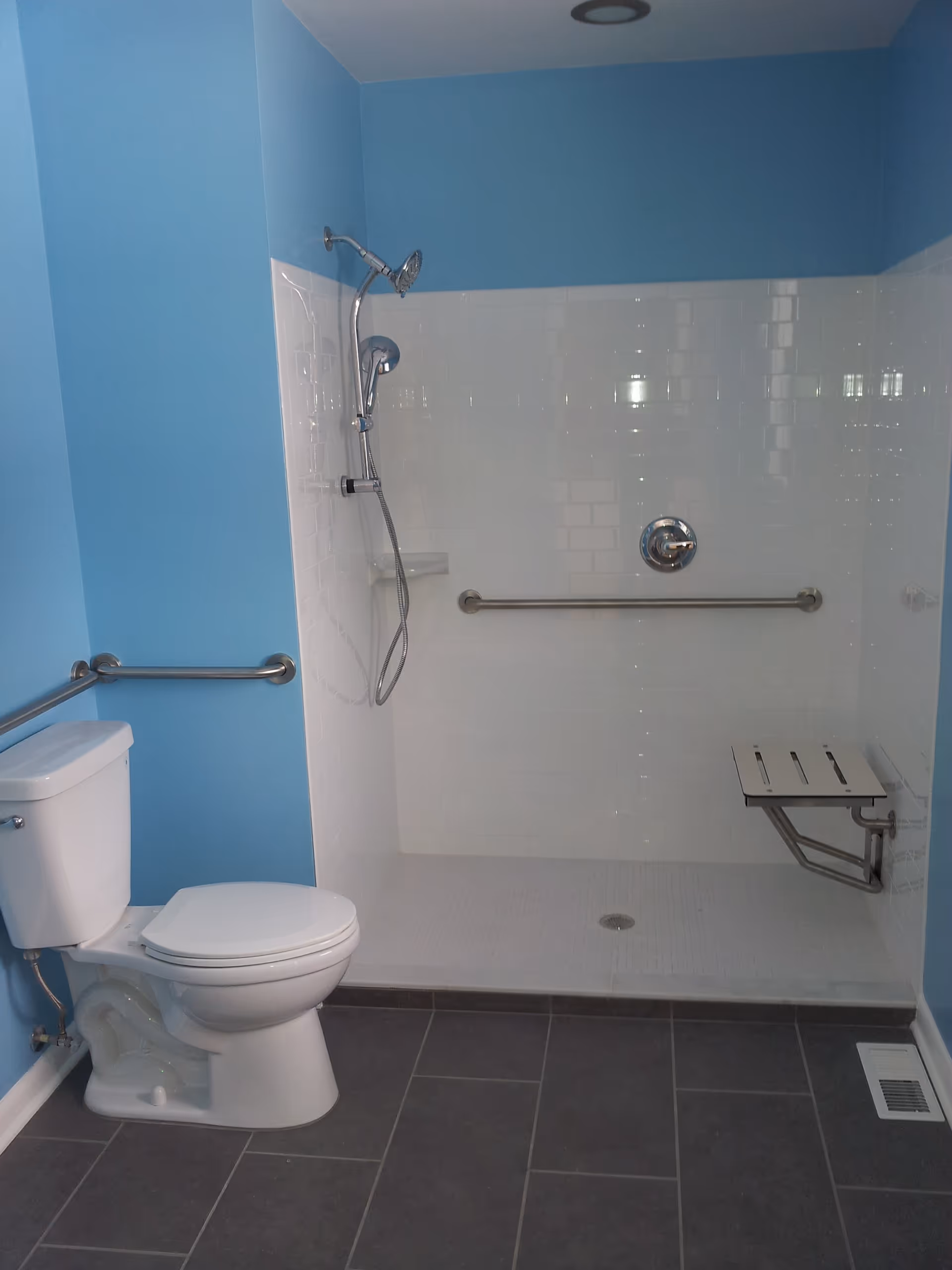 Bathroom with light blue walls featuring a white toilet and a walk-in shower with white tiled walls, a handheld showerhead, a horizontal grab bar, and a foldable shower seat. The floor is covered with dark gray tiles.