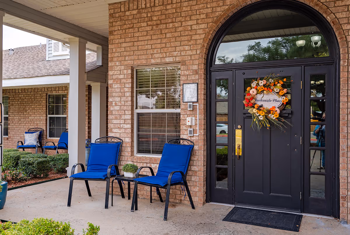 Front entrance of a brick building with a black door decorated with a colorful floral wreath. Two blue cushioned chairs and a small table with a plant are placed on the porch. Additional seating is visible in the background along with windows and landscaping.