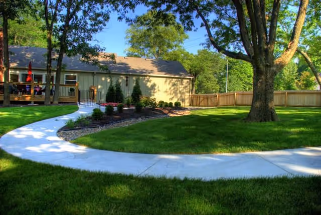 Curved concrete walkway through a landscaped green lawn with trees, a wooden fence, and a single-story building in the background.