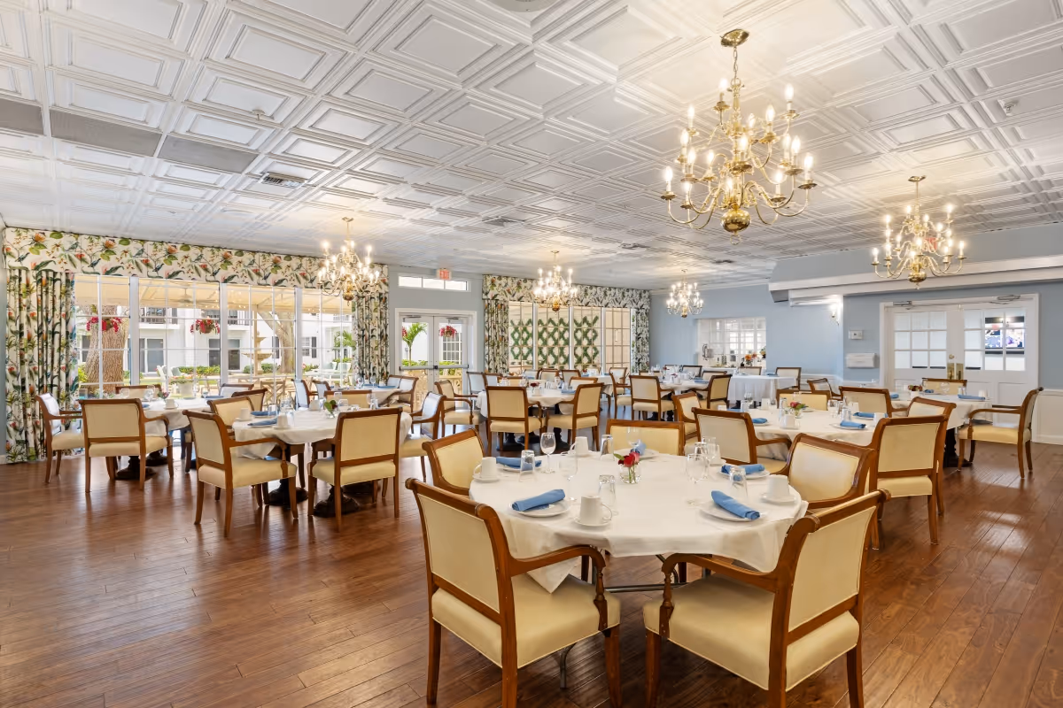 A spacious and elegant dining room with multiple round tables covered with white tablecloths, each set with blue napkins, glassware, and small floral centerpieces. The room features wooden flooring, large windows with floral curtains allowing natural light, and several ornate chandeliers hanging from a decorative white ceiling.