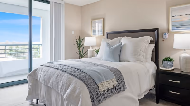 A neatly made bed with white linens, a gray throw blanket, and a light blue pillow in a bright bedroom. The room features a sliding glass door leading to a balcony with a view of trees and a clear sky. There are two bedside tables with lamps, a small plant, and framed artwork on the walls.
