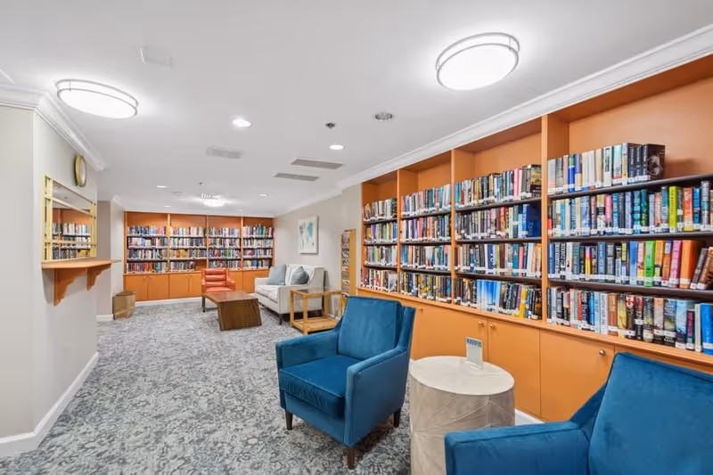 A cozy library room in a senior living facility featuring orange bookshelves filled with books, two blue armchairs with a small round table between them, a white sofa with cushions, an orange armchair, a wooden coffee table, a carpeted floor, and bright ceiling lights.