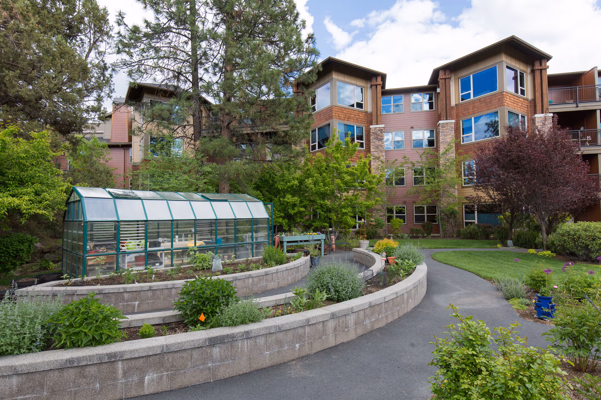 Courtyard with raised stone garden beds and a greenhouse in front of a multi-story senior living building.