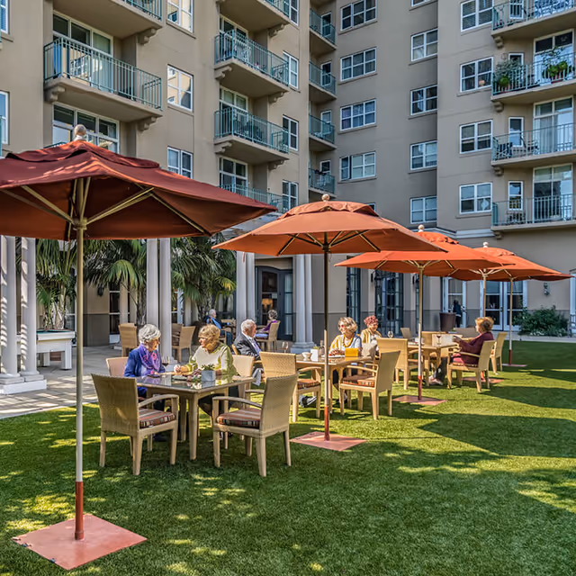 Several elderly people sitting at outdoor tables with large red umbrellas on a grassy area in front of a multi-story residential building, enjoying conversation and drinks.