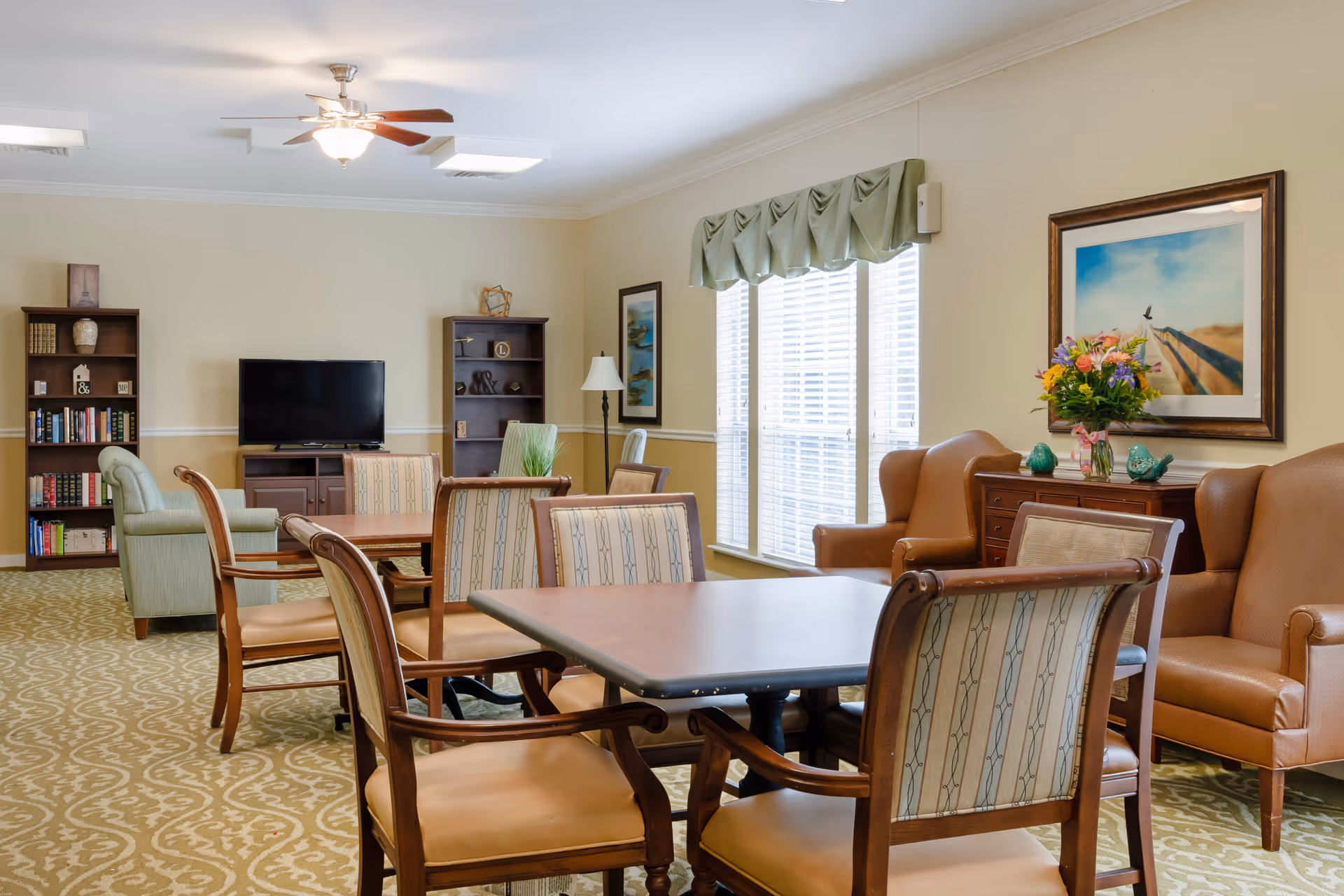 Bright senior living common room with tables and chairs, armchairs, bookshelves, and a TV by large windows.