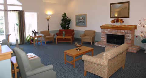 A cozy living room with multiple upholstered chairs and sofas arranged around a wooden coffee table and a brick fireplace. There is a tall floor lamp, a framed picture on the wall, a large mirror above the fireplace, and some plants near the windows letting in natural light.