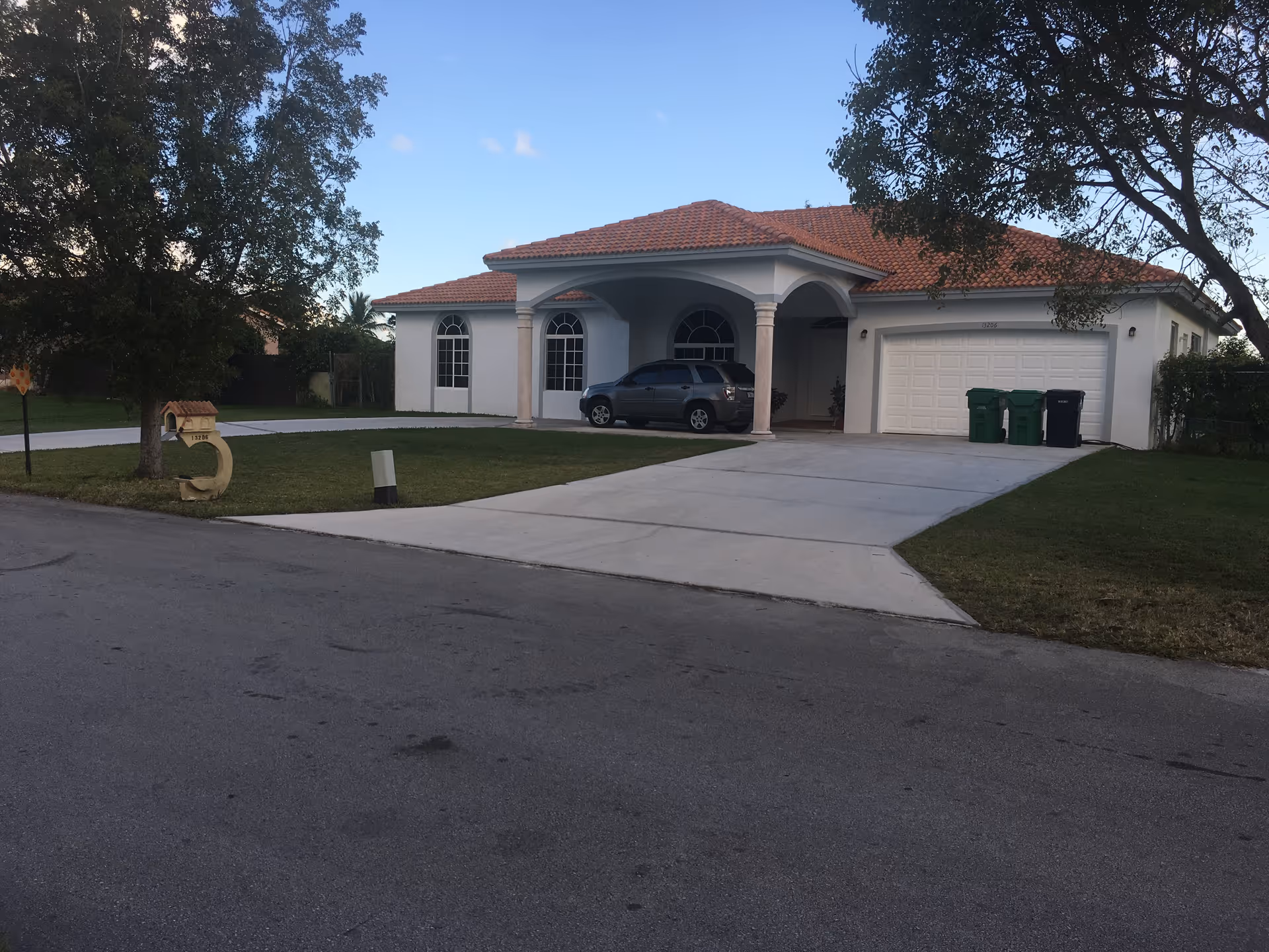 A single-story house with a red tiled roof and white exterior walls. There is a driveway leading to a garage with two green trash bins and one black trash bin beside it. A gray SUV is parked under a covered porch supported by columns. The house is surrounded by a lawn with trees on either side. The sky is clear and blue.