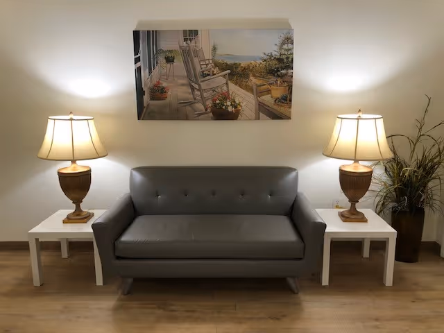 A small gray leather sofa is centered against a white wall, flanked by two white side tables each holding a table lamp with a beige shade. Above the sofa hangs a painting depicting a porch with rocking chairs and potted plants overlooking a scenic landscape. To the right of the sofa is a tall plant in a dark pot.