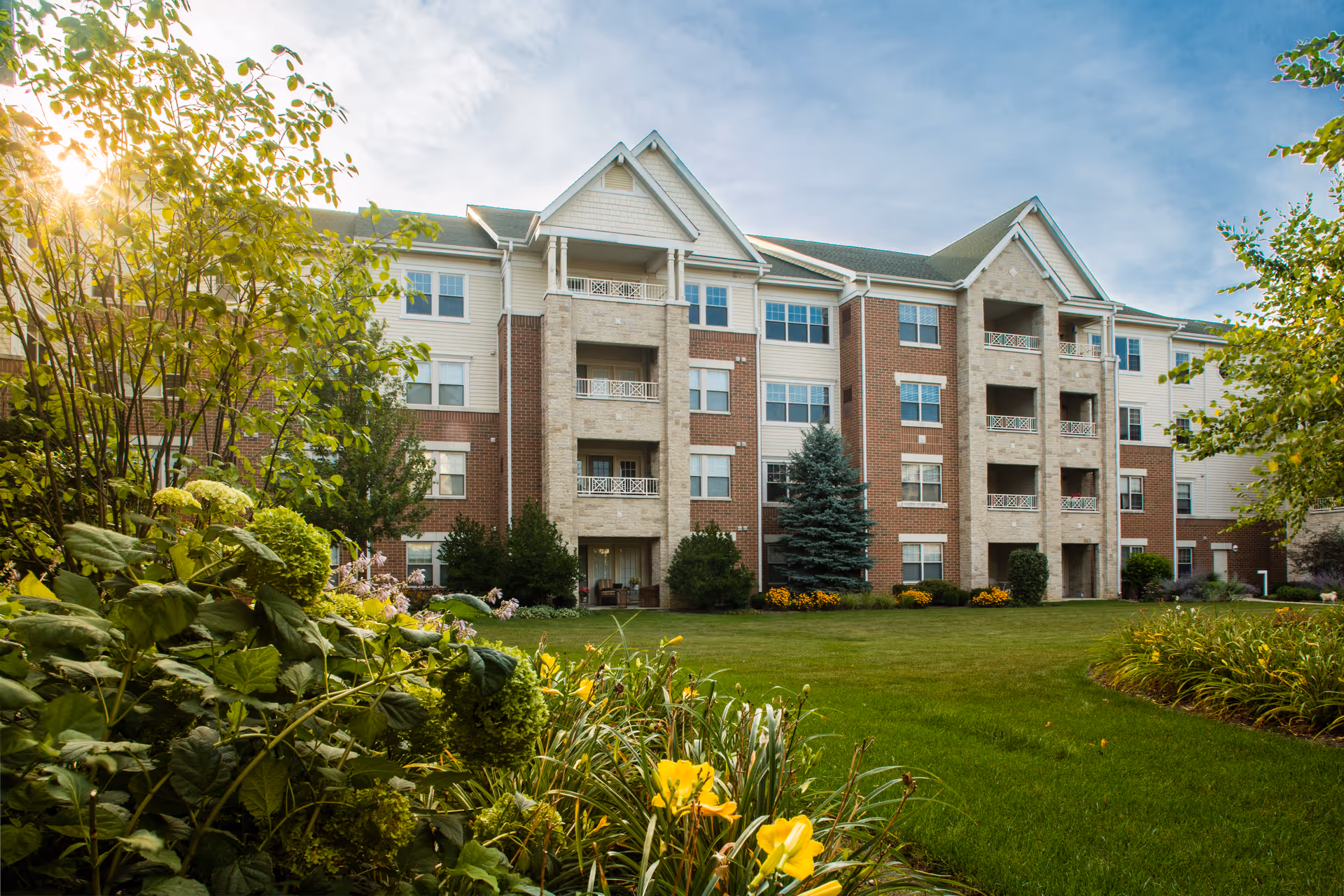 Front view of a multi-story brick and siding senior living building with balconies, lawn, and flowering landscaping.
