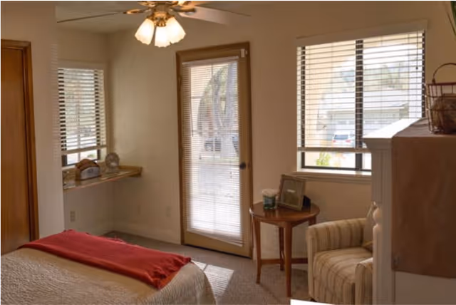 Sunlit bedroom with a bed topped by a red throw, an armchair, a round side table, and a glass-paned door and windows with blinds.