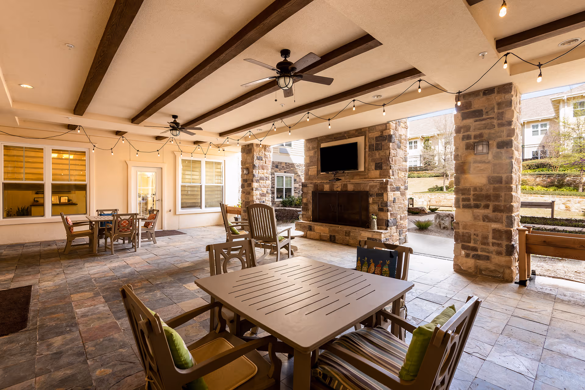 Covered outdoor patio with tables and chairs, ceiling fans, string lights, stone pillars, and a fireplace with a mounted TV.