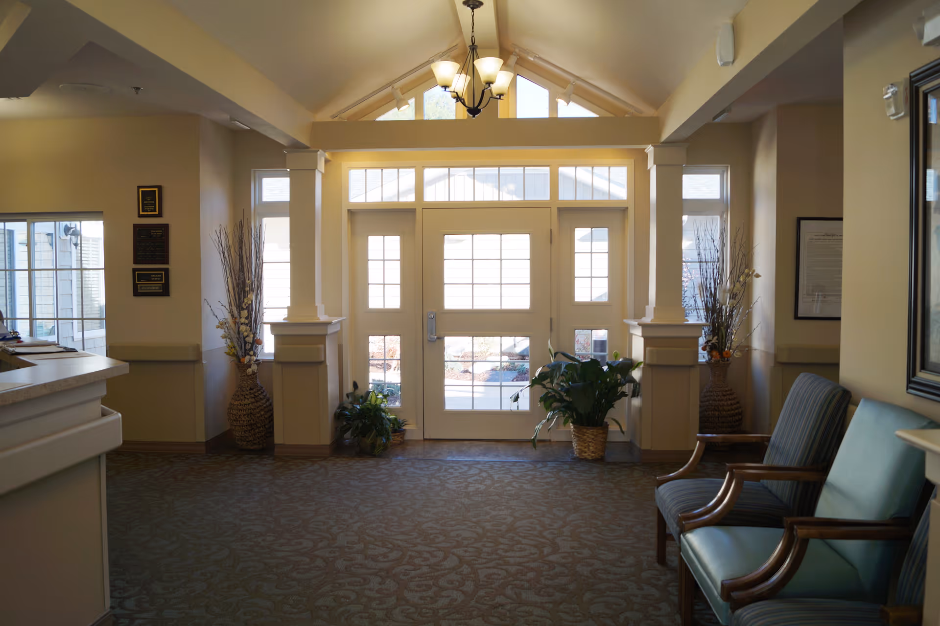 Sunlit lobby entryway with glass double doors, seating, decorative plants, and a reception counter.