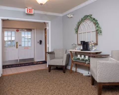 A cozy seating area in a senior living facility with two patterned armchairs, a wooden console table decorated with candles and greenery, and a decorative wall mirror shaped like a window. The area is carpeted and leads to a double door exit with glass panels.