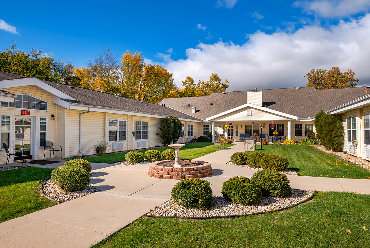 Outdoor courtyard area of Chandler Place senior living facility with a circular brick planter in the center, surrounded by neatly trimmed bushes and green grass. The building is a single-story structure with beige siding and multiple windows. There are chairs placed near the entrance doors, and the sky is partly cloudy with blue patches.