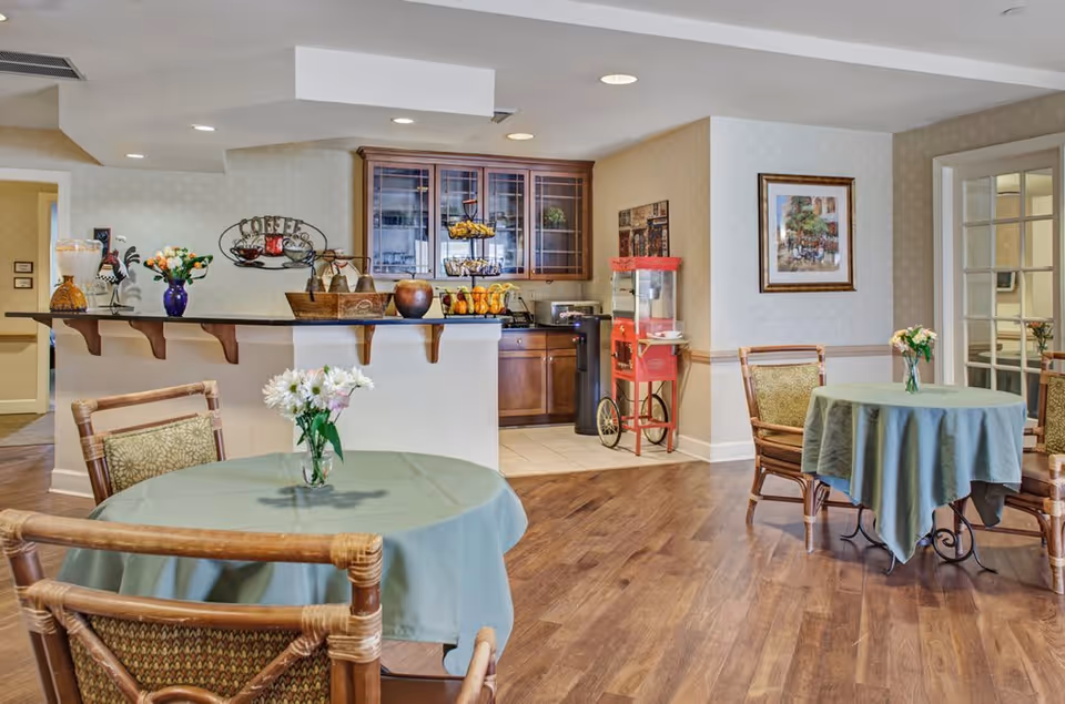 A cozy dining area with round tables covered in green tablecloths and decorated with small flower vases. The room features wooden chairs with patterned cushions, a wooden floor, and a counter with a coffee station and snacks. There is a red popcorn machine near the wall, framed artwork, and glass-paneled doors leading to another room.