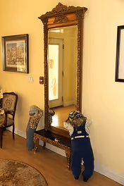 A tall ornate wooden mirror with intricate carvings stands against a beige wall. In front of the mirror, there are two child-sized mannequins dressed in overalls and hats. To the left, there is a wooden chair with a cushioned seat and a framed picture hanging on the wall above it. The floor is wooden, and part of a round rug is visible in the bottom left corner.