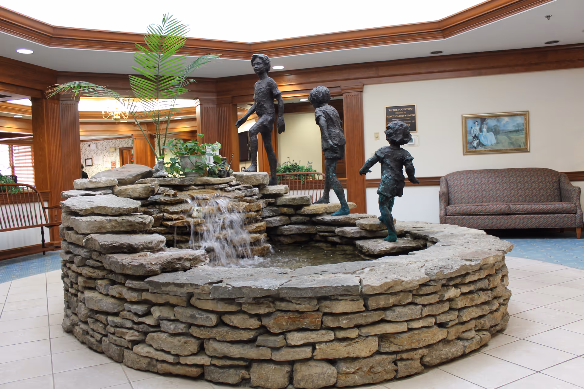 Indoor stone water fountain with three bronze statues of children playing, surrounded by wooden pillars and a seating area with a patterned couch and framed artwork on the wall in the background.