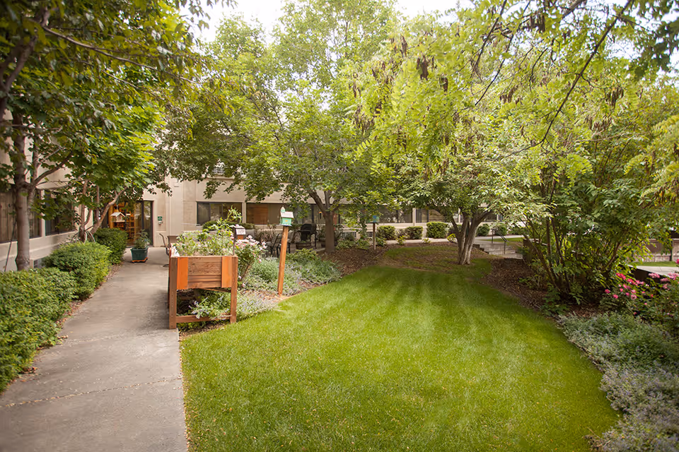 A lush outdoor garden area at Quail Ridge with a well-maintained green lawn, various trees, shrubs, and a concrete walkway. Raised garden beds with plants are visible along the path, and the building exterior with windows is seen in the background.