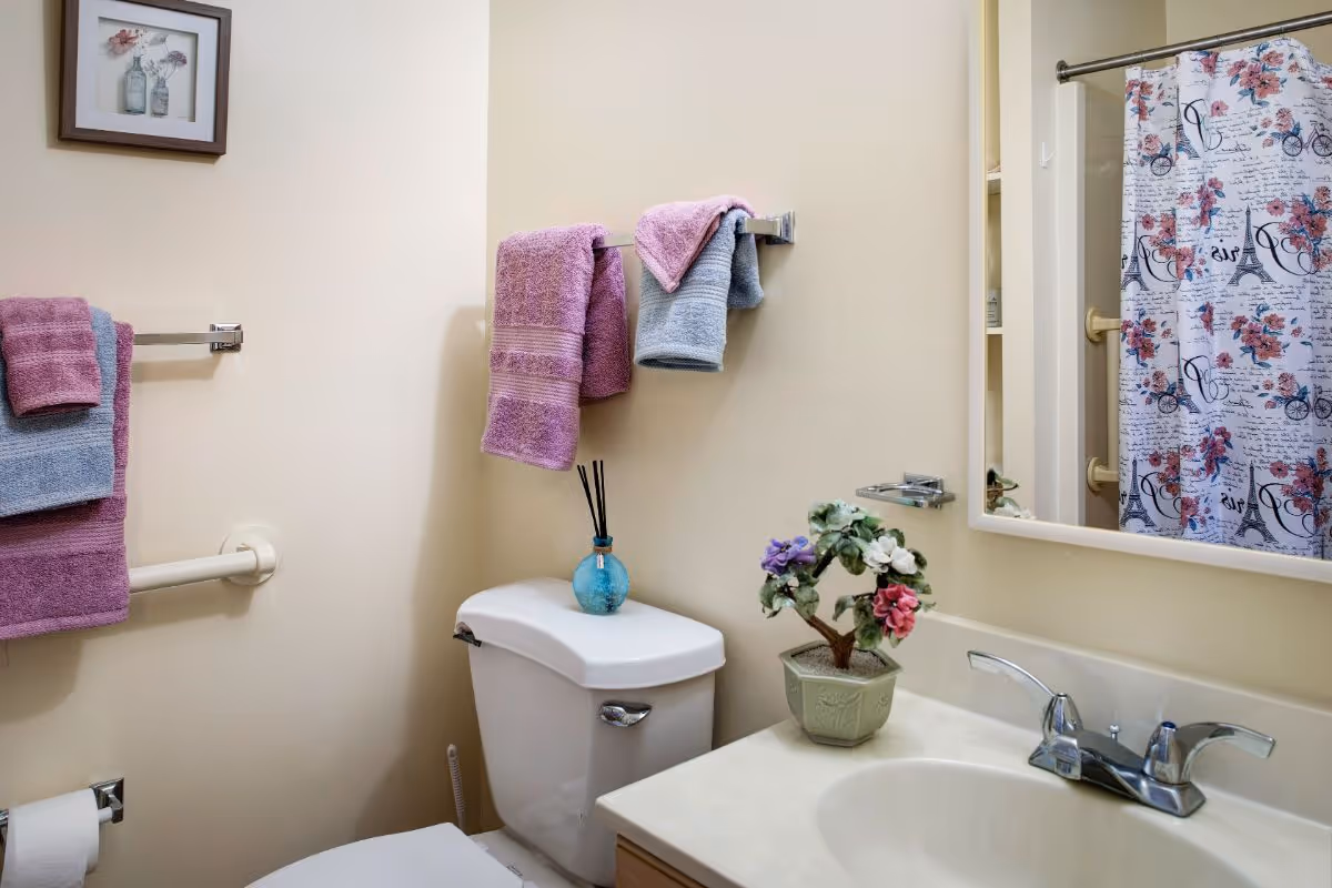 A small bathroom with a white toilet and a sink with a countertop. Above the toilet are two towel racks holding pink and blue towels. On the toilet tank is a small blue vase with reed diffuser sticks. Next to the sink is a small potted plant with colorful flowers. A mirror is mounted on the wall above the sink, reflecting a shower curtain with a floral and Eiffel Tower design. A framed picture hangs on the wall above the towel rack.