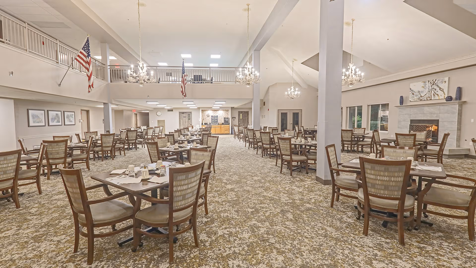 Spacious dining room with multiple wooden tables and chairs arranged neatly. The room features a high ceiling with chandeliers, American flags hanging from the upper balcony, a fireplace on the right wall, and large windows letting in natural light. The carpet has a patterned design and the walls are decorated with framed pictures.