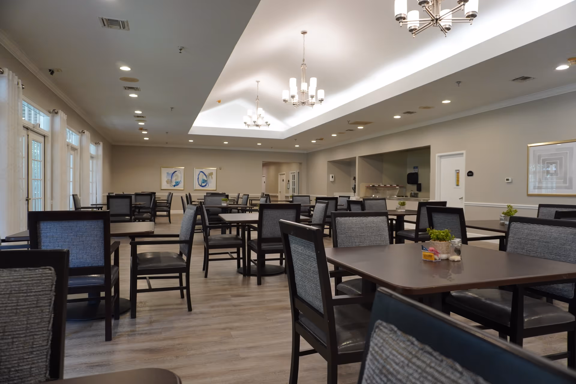Spacious dining room with multiple tables and chairs, chandeliers, and a serving area at the back.