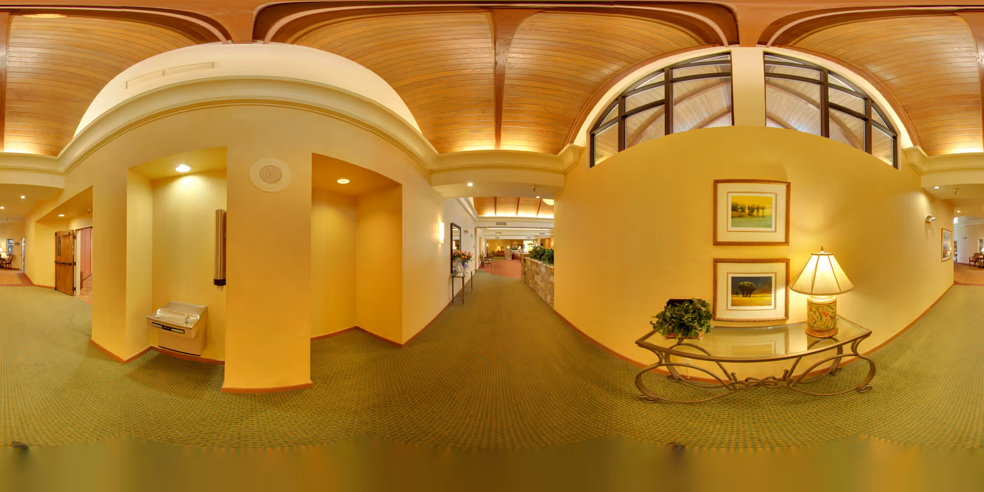 Warmly lit interior lobby hallway with an arched wooden ceiling, a glass console table with a lamp and framed art, and a recessed drinking fountain alcove.