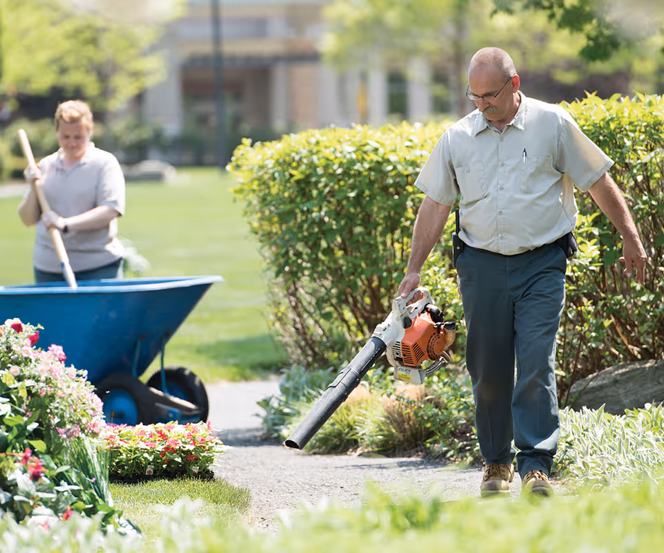 A man using a leaf blower on a garden path while a woman in the background pushes a blue wheelbarrow filled with gardening tools and plants, surrounded by greenery and flowers in an outdoor garden area.