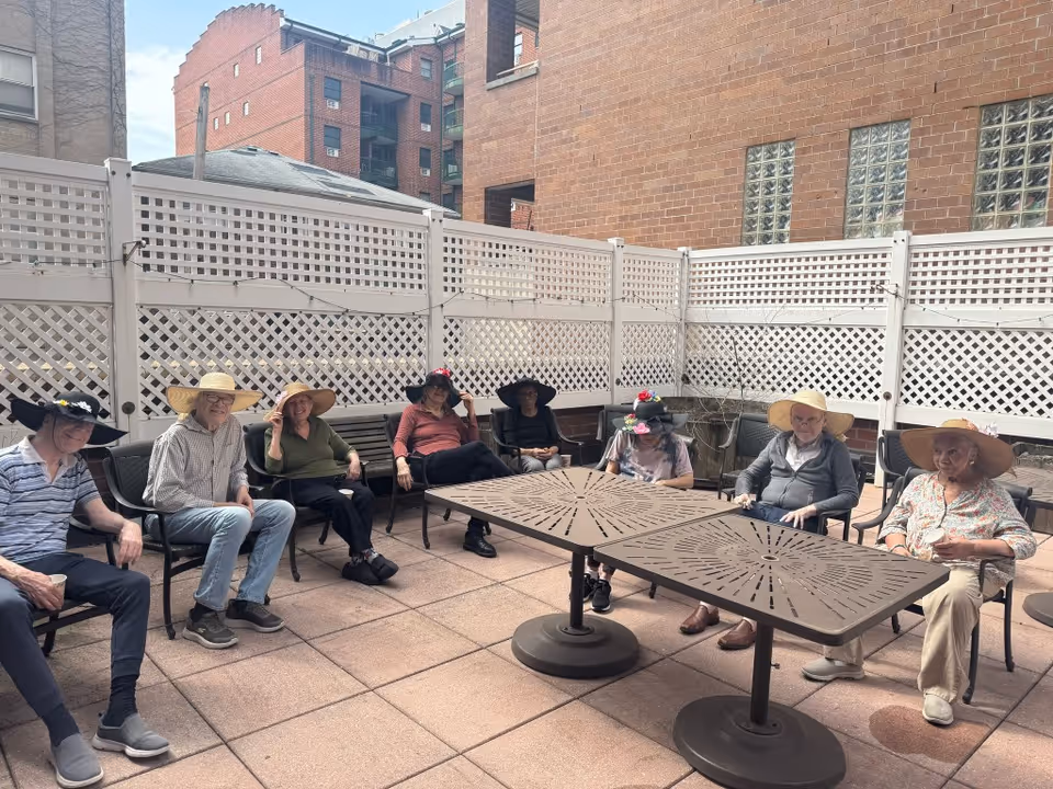 A group of elderly people sitting outdoors on chairs arranged in a semi-circle around two metal tables. They are wearing various hats and appear to be enjoying a sunny day on a patio enclosed by white lattice fencing and surrounded by brick buildings.
