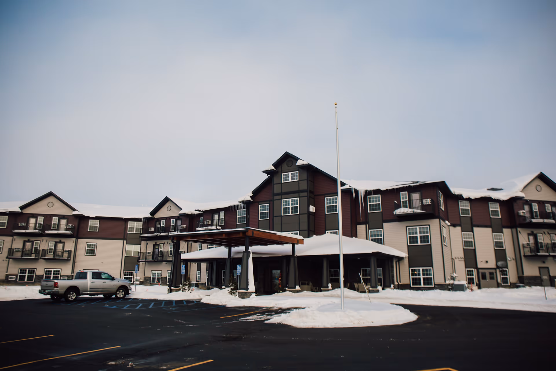 Front exterior of a multi-story senior living building with a covered entrance, flagpole, parked truck, and snow on the ground and roof.