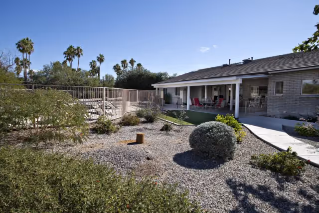 Outdoor view of a single-story building with a covered patio area furnished with tables and chairs. The foreground features a landscaped garden with gravel, bushes, and small plants. Palm trees and other greenery are visible in the background under a clear blue sky.