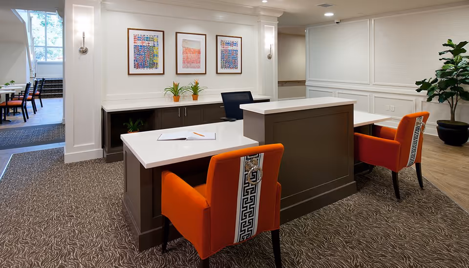 Reception desk area with two orange upholstered chairs, a white countertop desk, framed artwork on the wall, and potted plants.