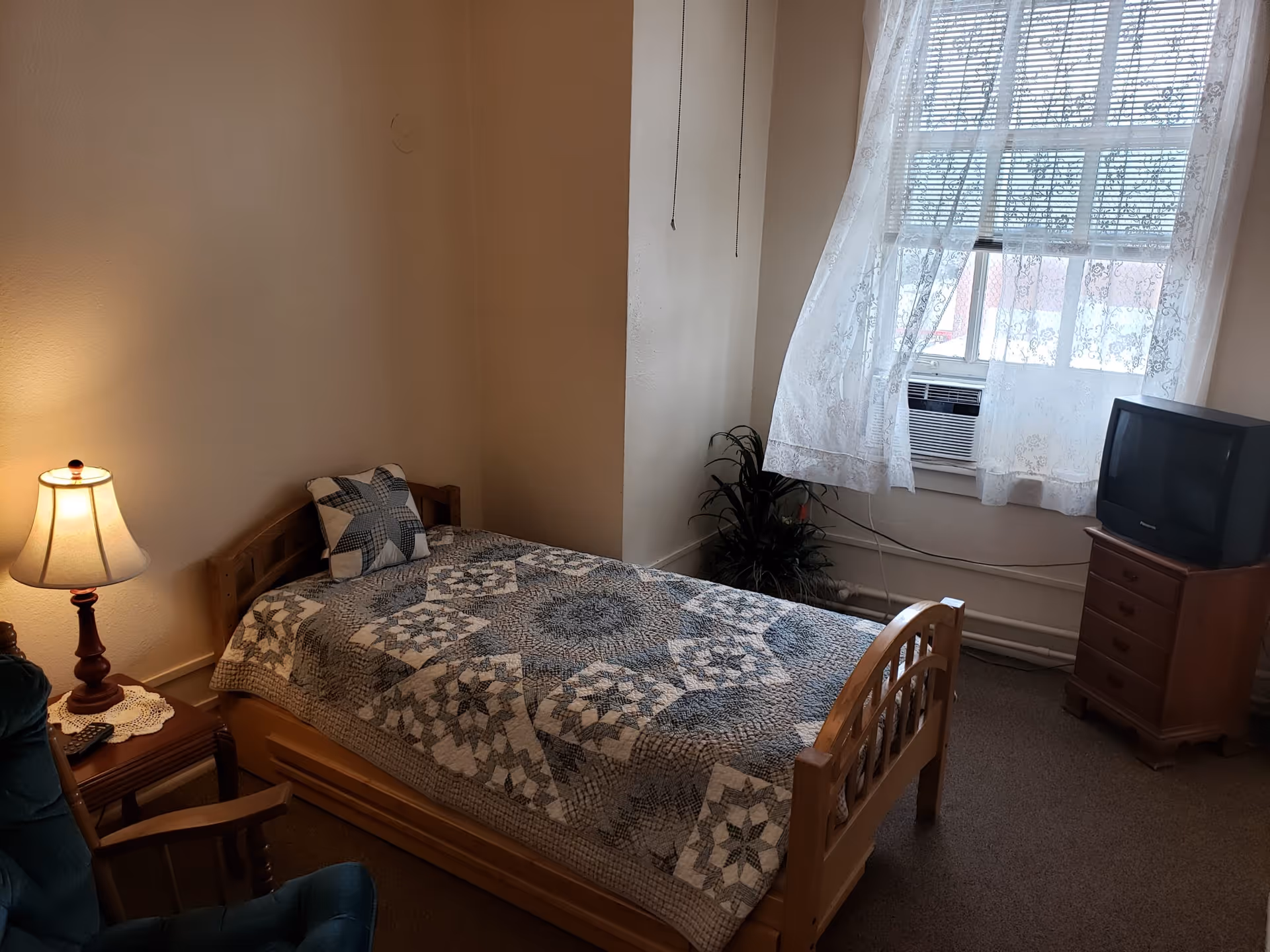A small bedroom with a single wooden bed covered with a patterned quilt and a matching pillow. Next to the bed is a wooden side table with a lamp and a remote control. A green upholstered chair is partially visible in the foreground. The room has a window with sheer white curtains and an air conditioning unit beneath it. A small wooden dresser with a TV on top is positioned near the window. There is a potted plant in the corner of the room.