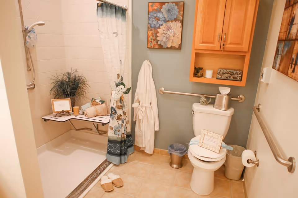 A bathroom with a walk-in shower featuring a floral shower curtain and a fold-down bench with towels and decorative items. A white bathrobe hangs on the wall next to the shower. The toilet has a tissue box on top and a framed note on the seat. There is a small trash can and a wastebasket beside the toilet. Above the toilet is a wooden cabinet with decorative items, and a floral painting hangs on the wall. The bathroom has beige tiled flooring and grab bars for accessibility.