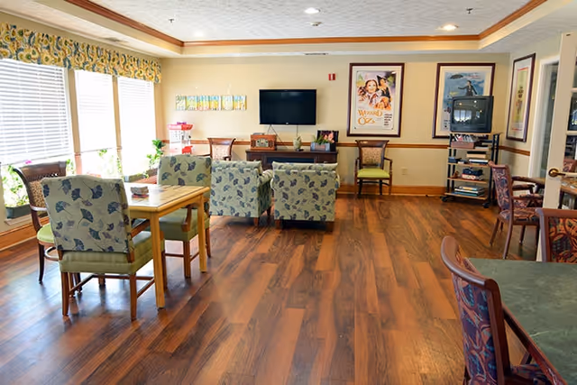A bright and spacious common area in a senior living facility with wooden floors, several tables and chairs, patterned armchairs, a flat screen TV mounted on the wall, and framed artwork including a Wizard of Oz poster. Large windows with floral valances let in natural light.