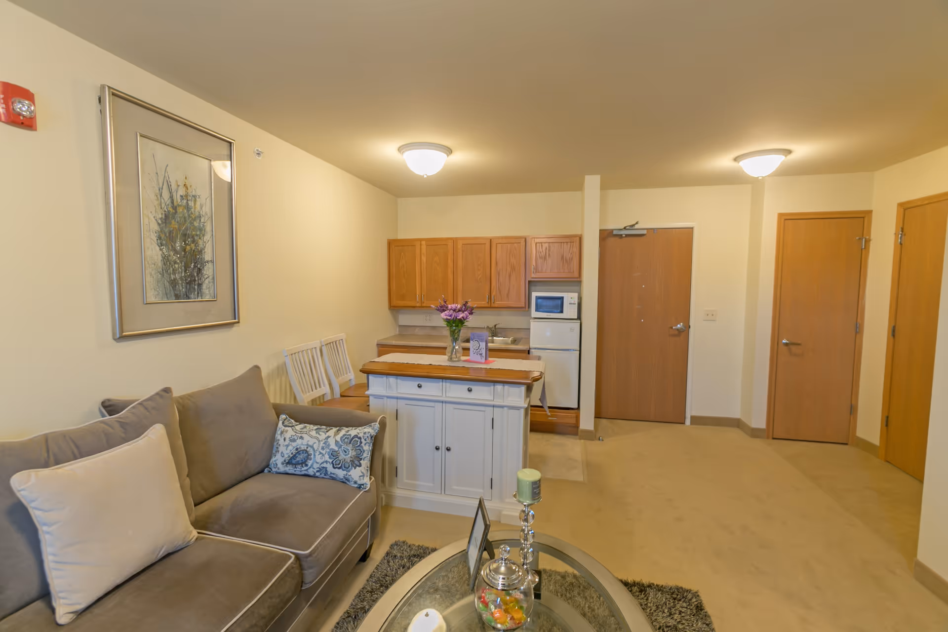 Interior view of a senior living facility apartment showing a small living area with a gray couch adorned with pillows, a glass coffee table with decorative items, a kitchen area with wooden cabinets, a microwave, and a refrigerator. There are two white chairs and a small island with a vase of flowers. The walls are light-colored, and there are three wooden doors visible in the background.