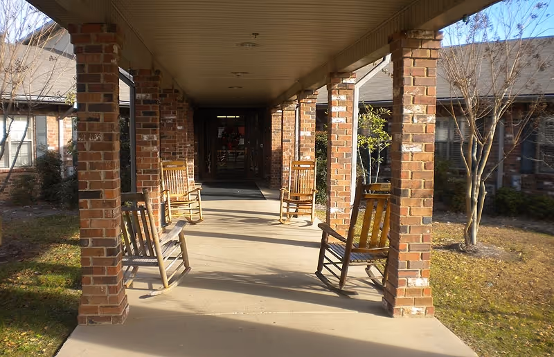 Covered outdoor walkway with brick pillars and several wooden rocking chairs placed along the sides, leading to the entrance of a building with glass doors. There are leafless trees and some shrubs on either side of the walkway.