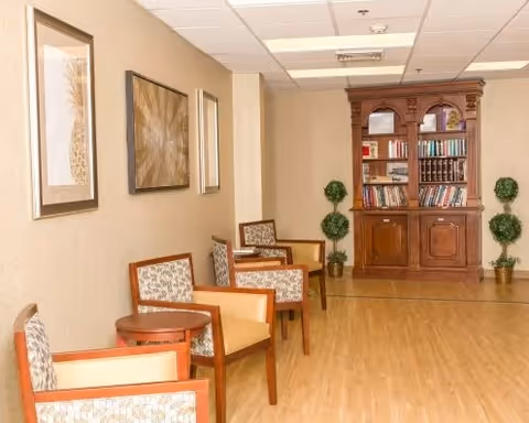 A hallway or common area with wooden flooring, beige walls, and ceiling tiles. There are several wooden chairs with patterned cushions and small round wooden tables along the left wall. On the far end, there is a wooden bookshelf filled with books and decorative items, flanked by two small potted topiary plants. Two framed artworks hang on the left wall.