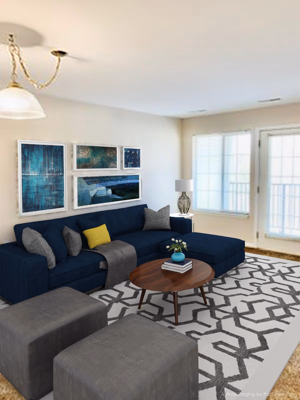 A modern living room with a dark blue sectional sofa adorned with gray and yellow pillows, a wooden coffee table with books and a small plant, two gray ottomans, a patterned area rug, a side table with a decorative lamp, and abstract blue artwork on the wall. Large windows and a glass door allow natural light into the room.