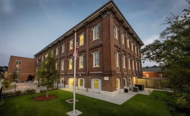 Exterior view of a three-story brick building with multiple windows illuminated from inside during dusk. There is a flagpole with an American flag in front of the building, surrounded by a well-maintained lawn and trees. The sky is partly cloudy.