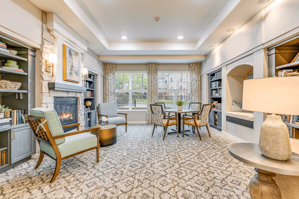 Well-lit senior living common room with a fireplace, armchairs, bookshelves, and a small table by a large window.