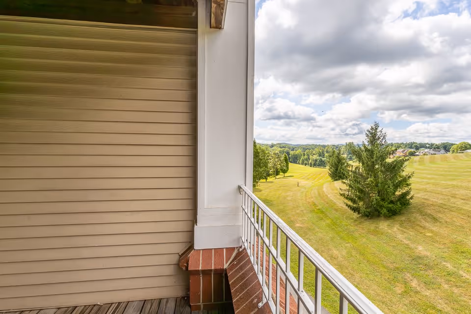 View from a balcony with a white railing overlooking a large grassy field with scattered trees under a partly cloudy sky. The balcony has a wooden floor and beige siding on one side.