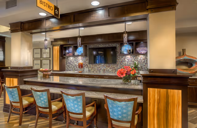 Interior view of a bistro area in an assisted living facility featuring a long granite countertop with four wooden chairs that have blue patterned backs and yellow seats. Behind the counter is a mosaic tile backsplash with a mounted flat-screen TV, shelves with decorative items, and three hanging pendant lights with blue shades. A small bouquet of red flowers is placed on the counter. The space has warm wood paneling and flooring, with a sign overhead that reads 'BISTRO'.