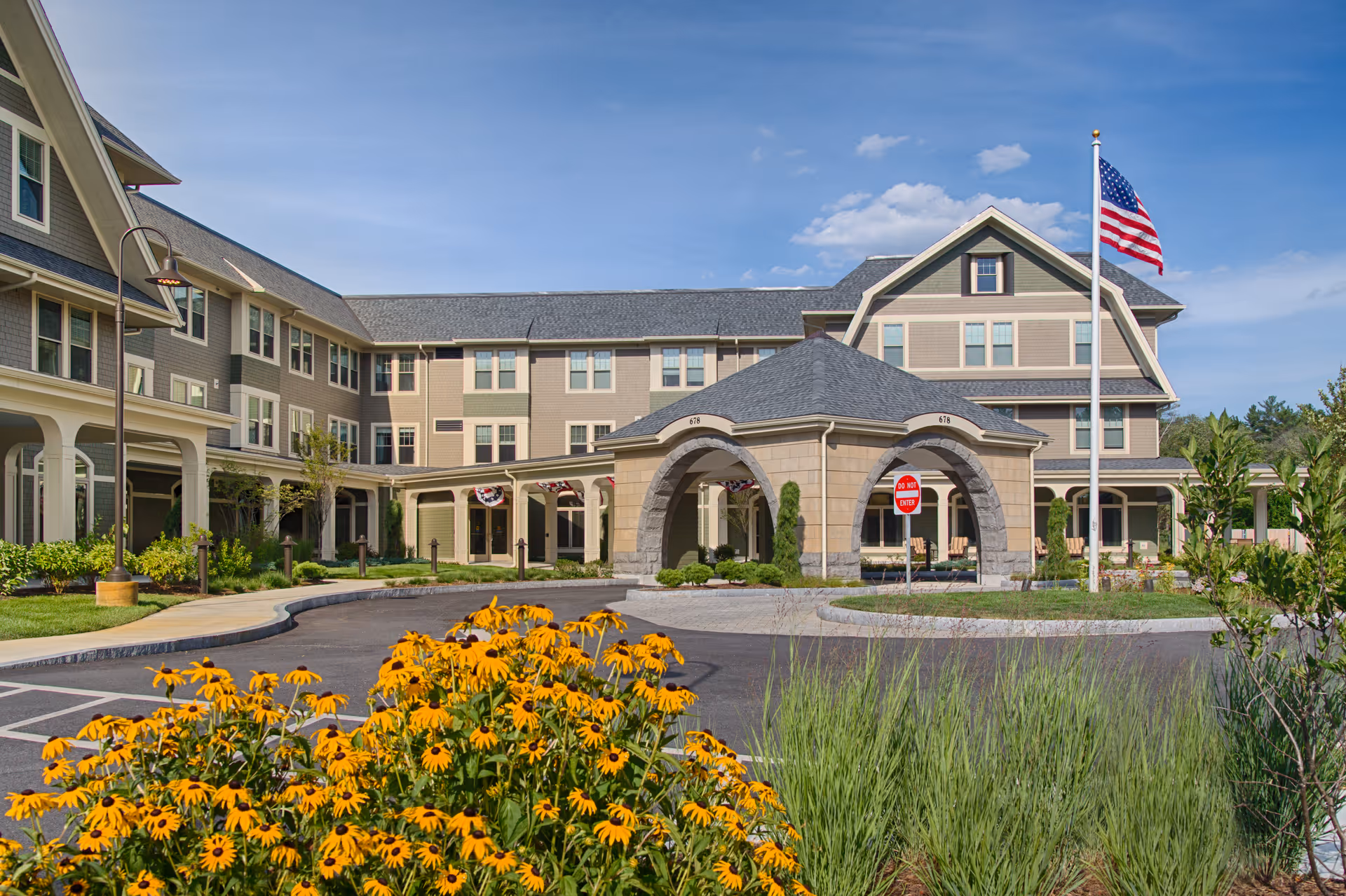 Front entrance of a multi-story senior living building with an arched porte-cochere, landscaping and an American flag.