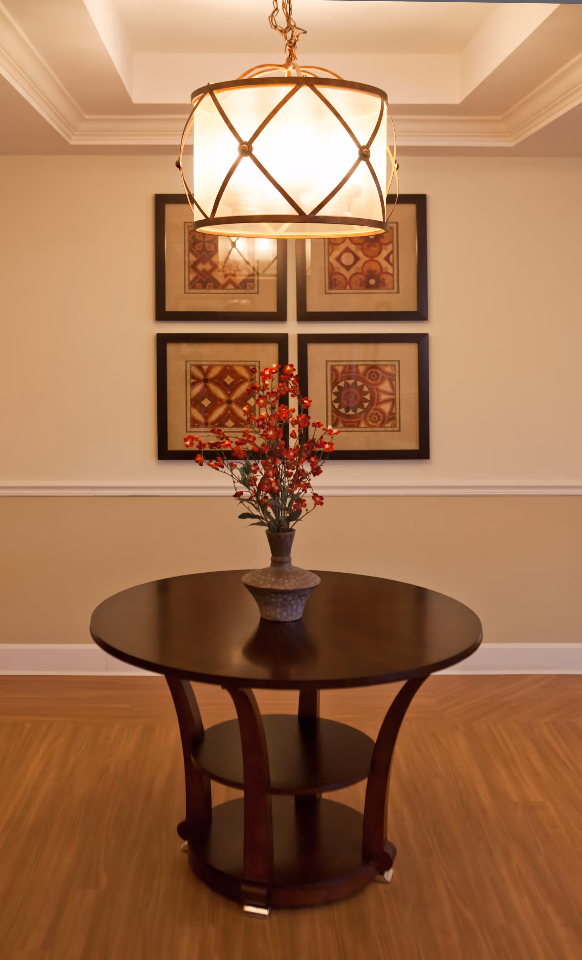 A round wooden table with a vase of red flowers on top, positioned in front of a wall with four framed decorative art pieces arranged in a square. Above the table hangs a large, round light fixture with a crisscross pattern.