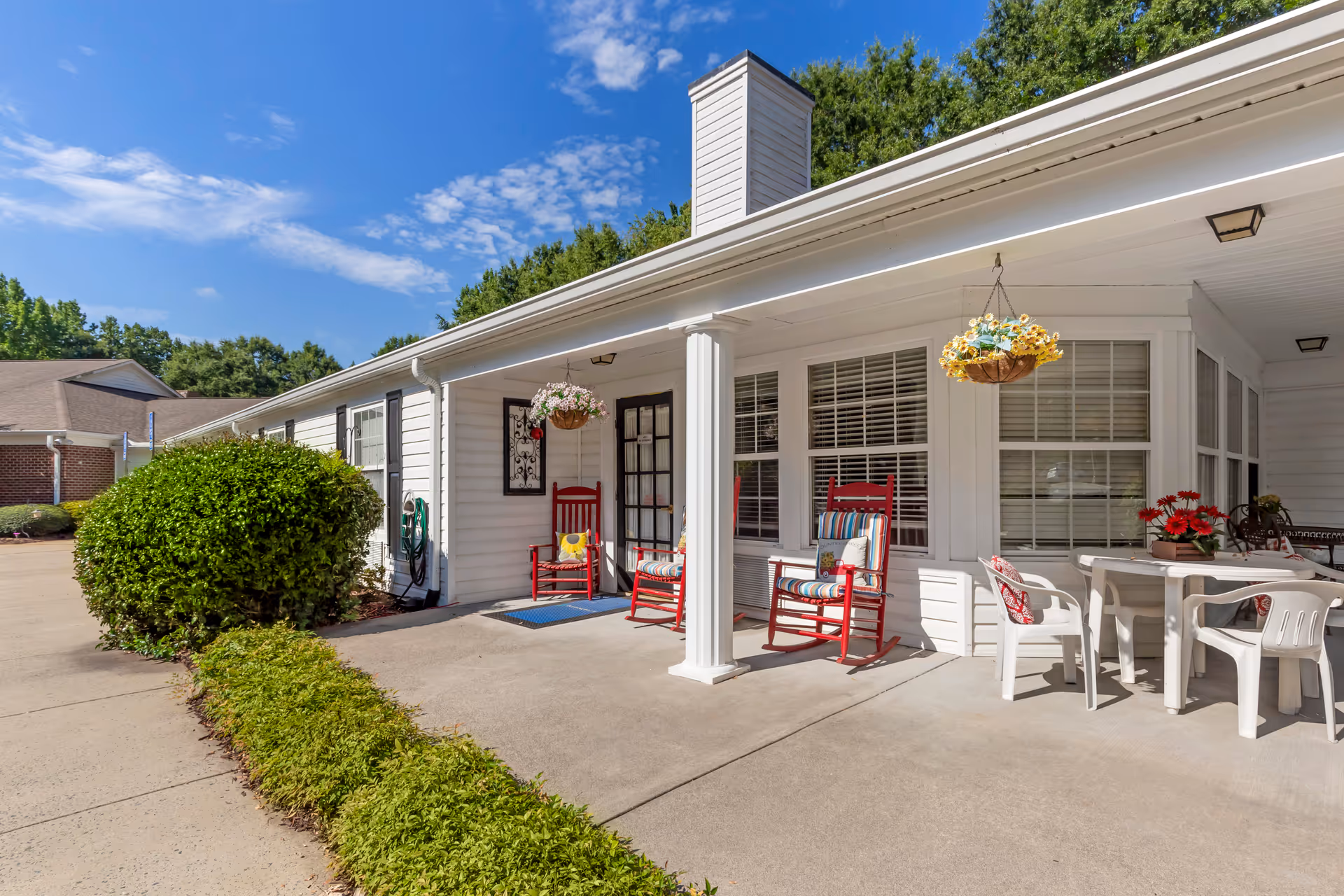 Outdoor patio area of a senior living facility with white siding walls, a covered porch supported by a white column, red rocking chairs with cushions, a white table with white chairs, hanging flower baskets, and green bushes along the concrete walkway under a blue sky with scattered clouds.