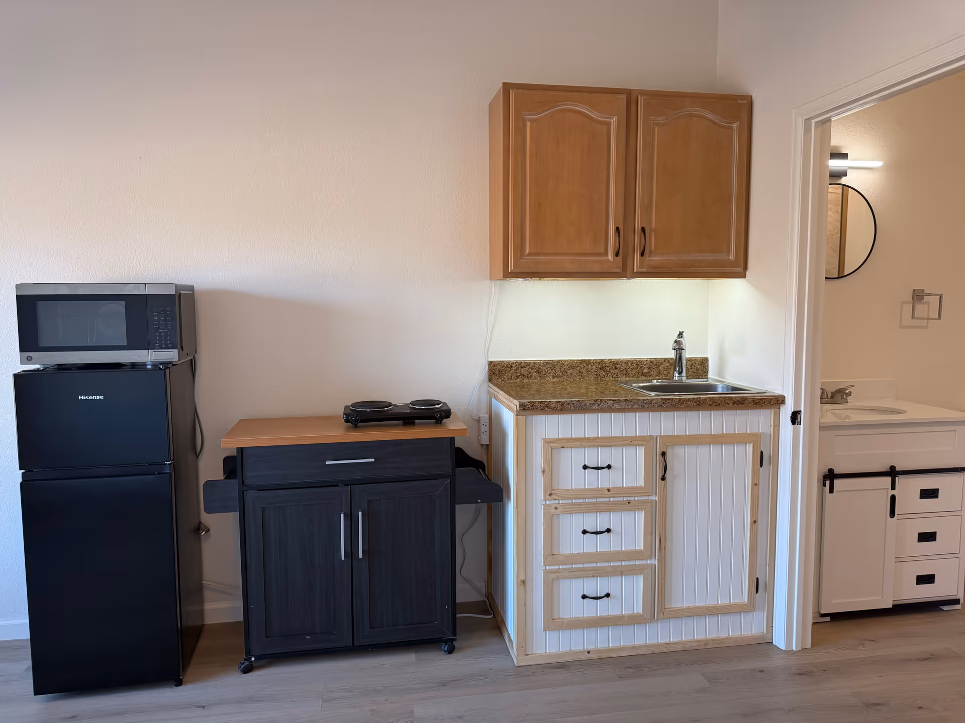 A small kitchenette area featuring a black Hisense refrigerator with a microwave on top, a wooden cart with a double hot plate, and a white cabinet with a brown countertop and a sink. To the right, there is a doorway leading to a bathroom with a white vanity and a round mirror.