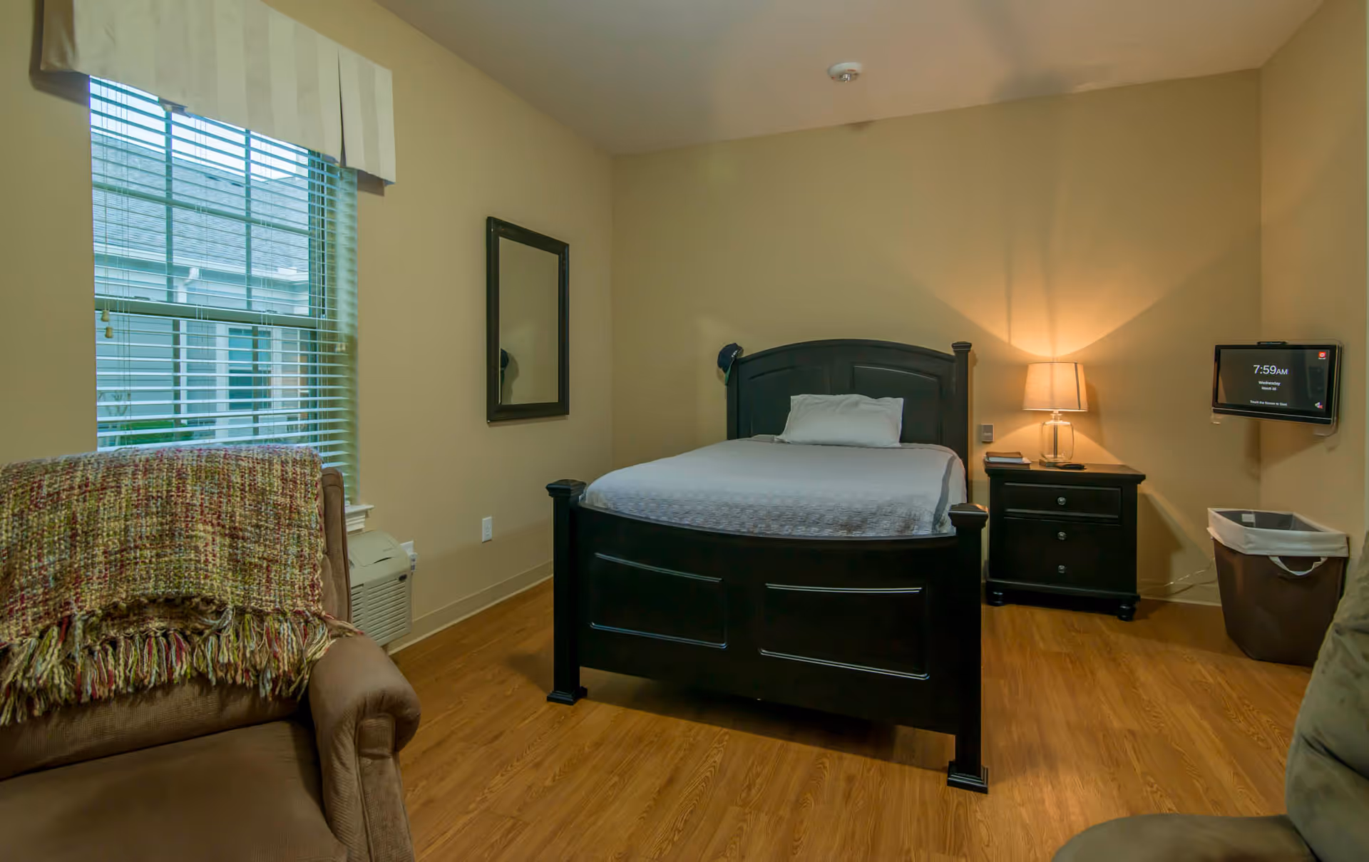 A cozy bedroom with a dark wooden bed frame and white bedding, a nightstand with a lamp, a wall-mounted TV, a window with blinds, a brown armchair with a multicolored throw blanket, and wooden flooring.