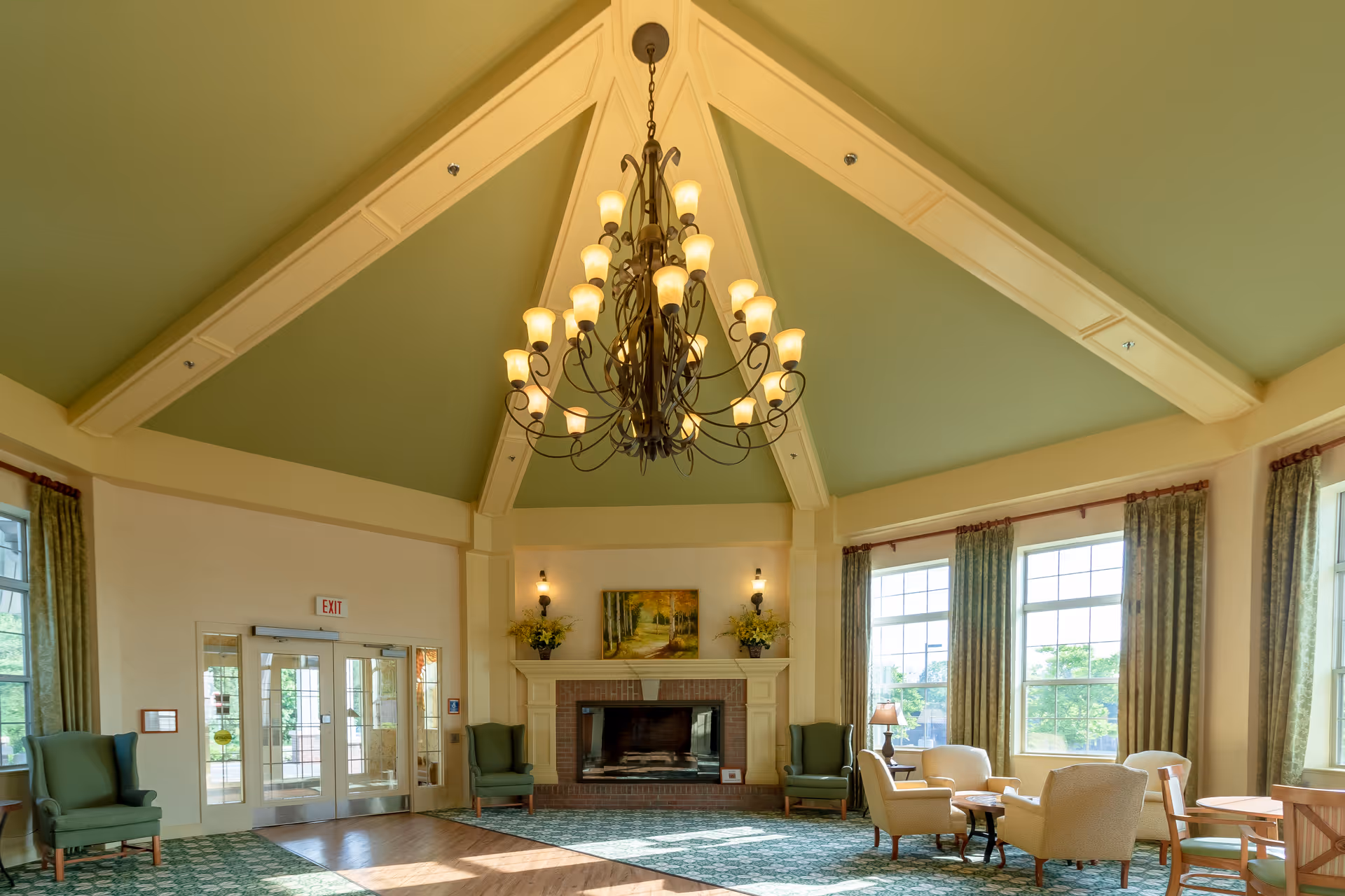 A spacious and well-lit common area with a high vaulted ceiling painted green and cream beams. A large ornate chandelier hangs from the ceiling. The room features a fireplace with a painting above it, flanked by two wall sconces and flower arrangements. There are several comfortable chairs and small tables arranged near large windows with green curtains, allowing natural light to fill the room. Double glass doors are visible on one side.