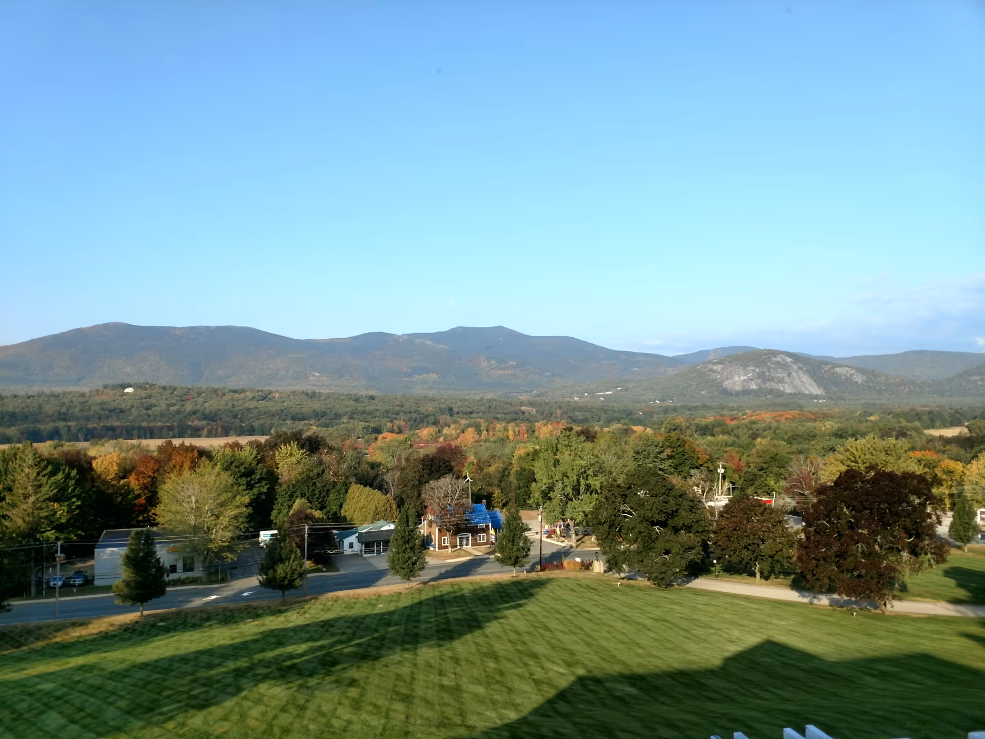 A scenic outdoor view showing a large green lawn in the foreground, a road lined with trees and small buildings, and a backdrop of forested hills and mountains under a clear blue sky.