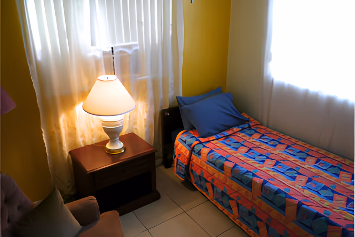 A small bedroom with a single bed covered in a colorful patterned bedspread and a blue pillow. Next to the bed is a wooden nightstand with a white lamp on it. The room has yellow walls and two windows with white curtains letting in natural light. A cushioned chair is partially visible in the corner.